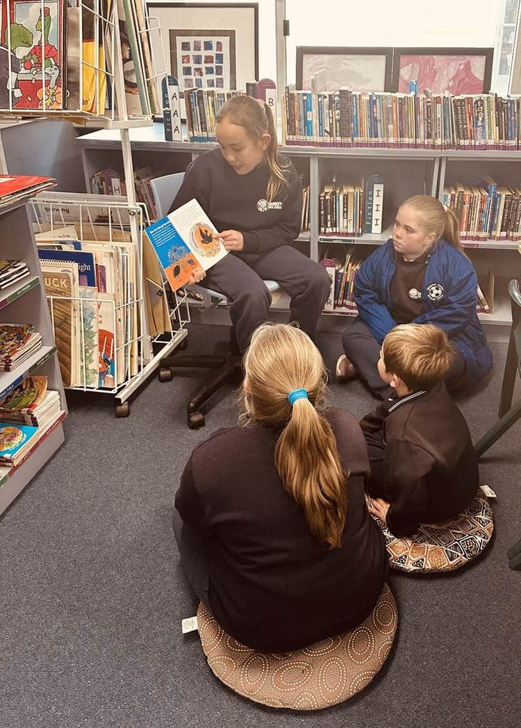 A primary student reading a book to 3 other students in the school library