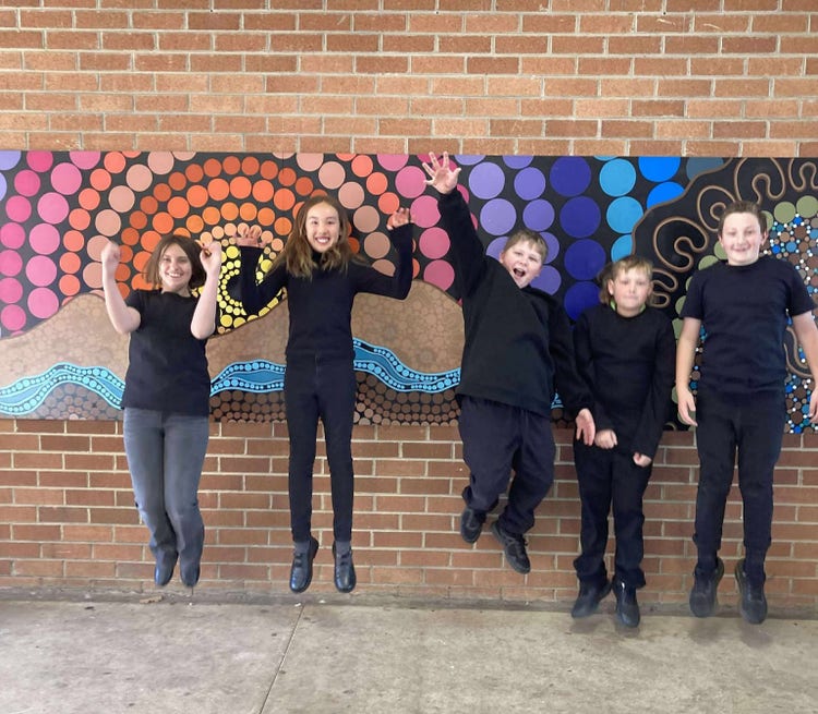 5 students from the Tournament of Minds team dressed in black, jumping in front of an Aboriginal artwork hung on a brick wall.