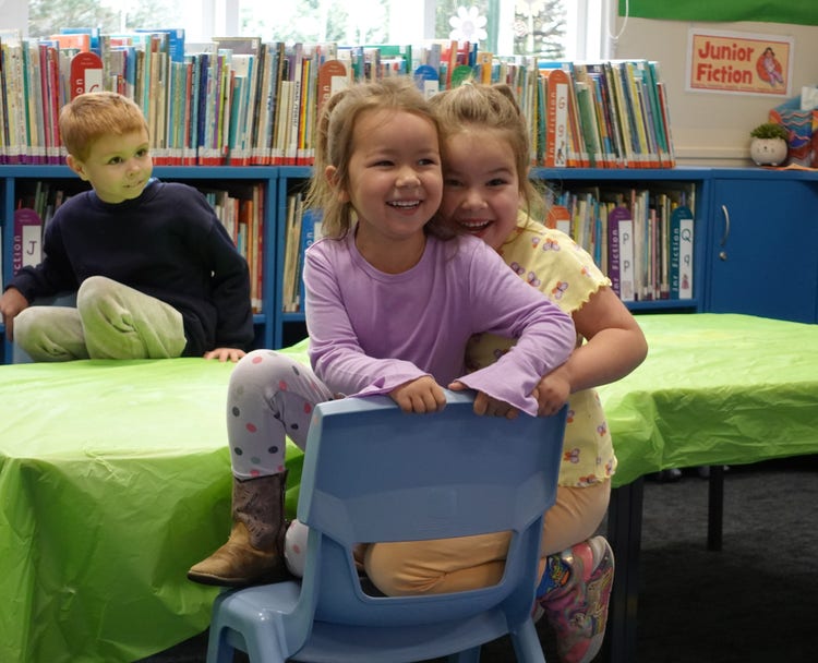 2 children sitting on a chair smiling. another child in the background also sitting on a chair. Bookshelves in the background