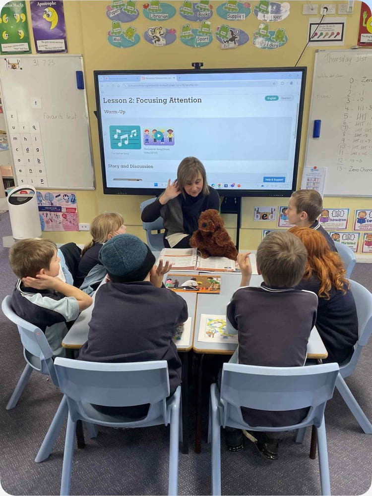 Trunkey Public School students sitting around a table with SLSO Mrs Brown leading a Second Step lesson. Mrs Brown has her hand to her ear and is holding a dog puppet.