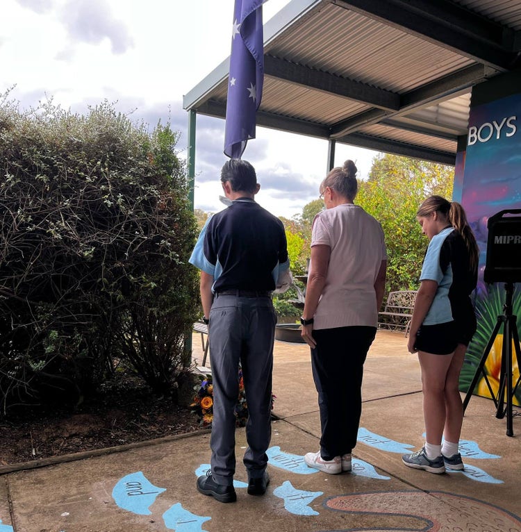 Two students and a teacher standing with heads bowed in front of the Australian flag at half mast after laying a wreath at the base.